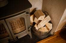 Load image into Gallery viewer, Firewood in  a basket next to a woodburning stove.
